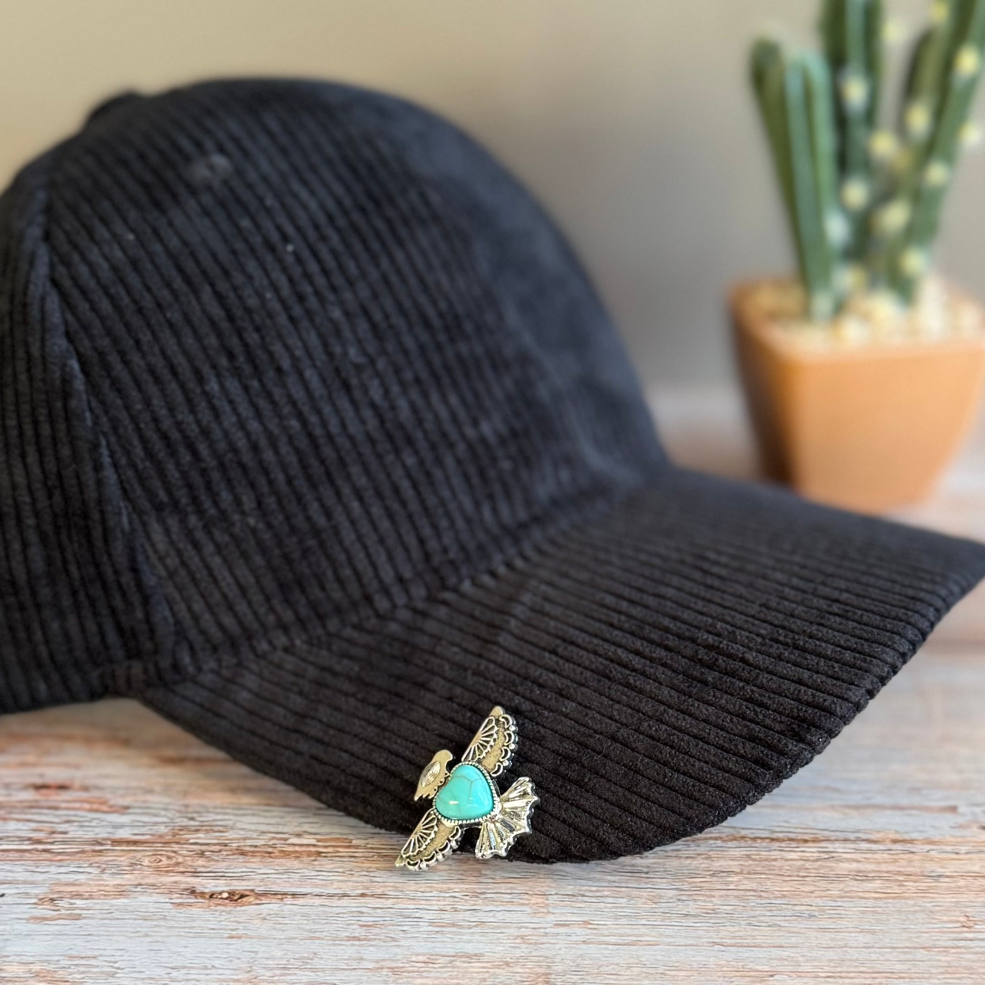 Black corduroy cap with a decorative brooch featuring a turquoise stone and gold accents on a wooden surface.