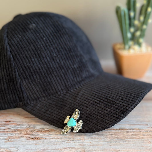 Black corduroy cap with a decorative brooch featuring a turquoise stone and gold accents on a wooden surface.