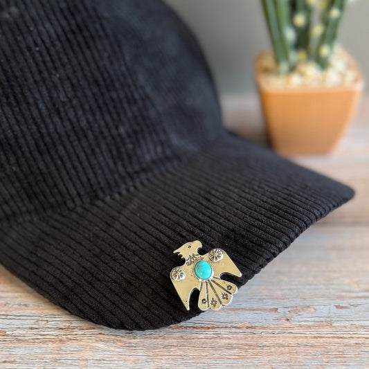 Black corduroy hat with a gold and turquoise brooch on a wooden surface with a cactus in the background.