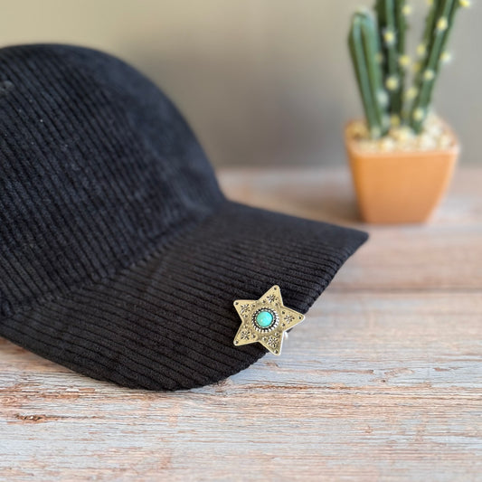 Black cap with a star-shaped brooch featuring a turquoise stone on a wooden surface with a blurred cactus plant in the background.