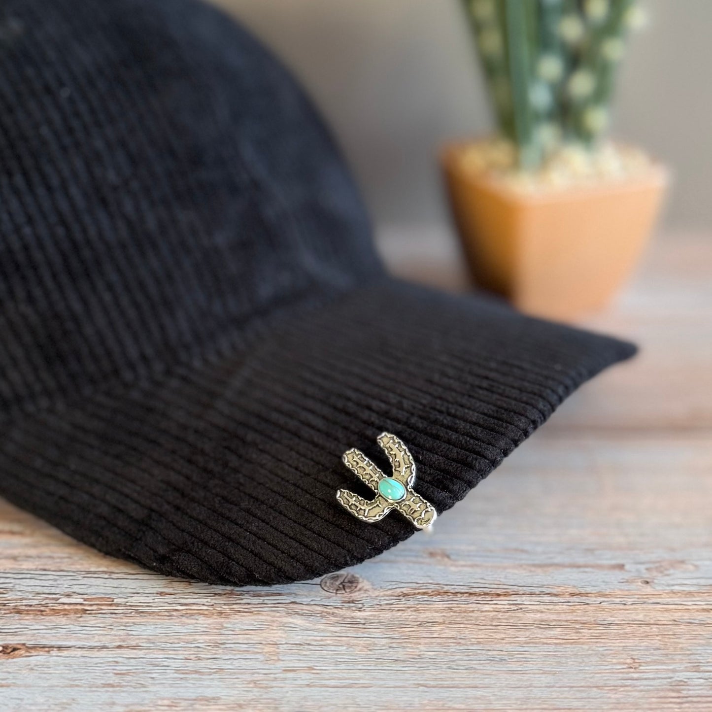 Black cap with a decorative cactus brooch on a wooden surface, with a blurred plant in the background.