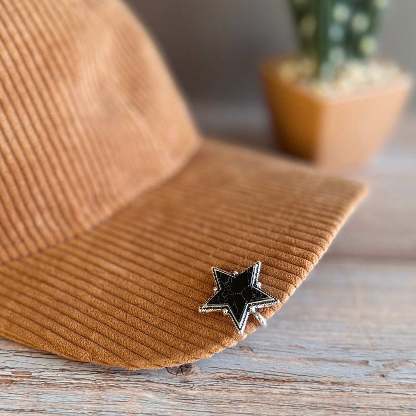Brown corduroy hat with a black star pin on a wooden surface, blurred plant in the background