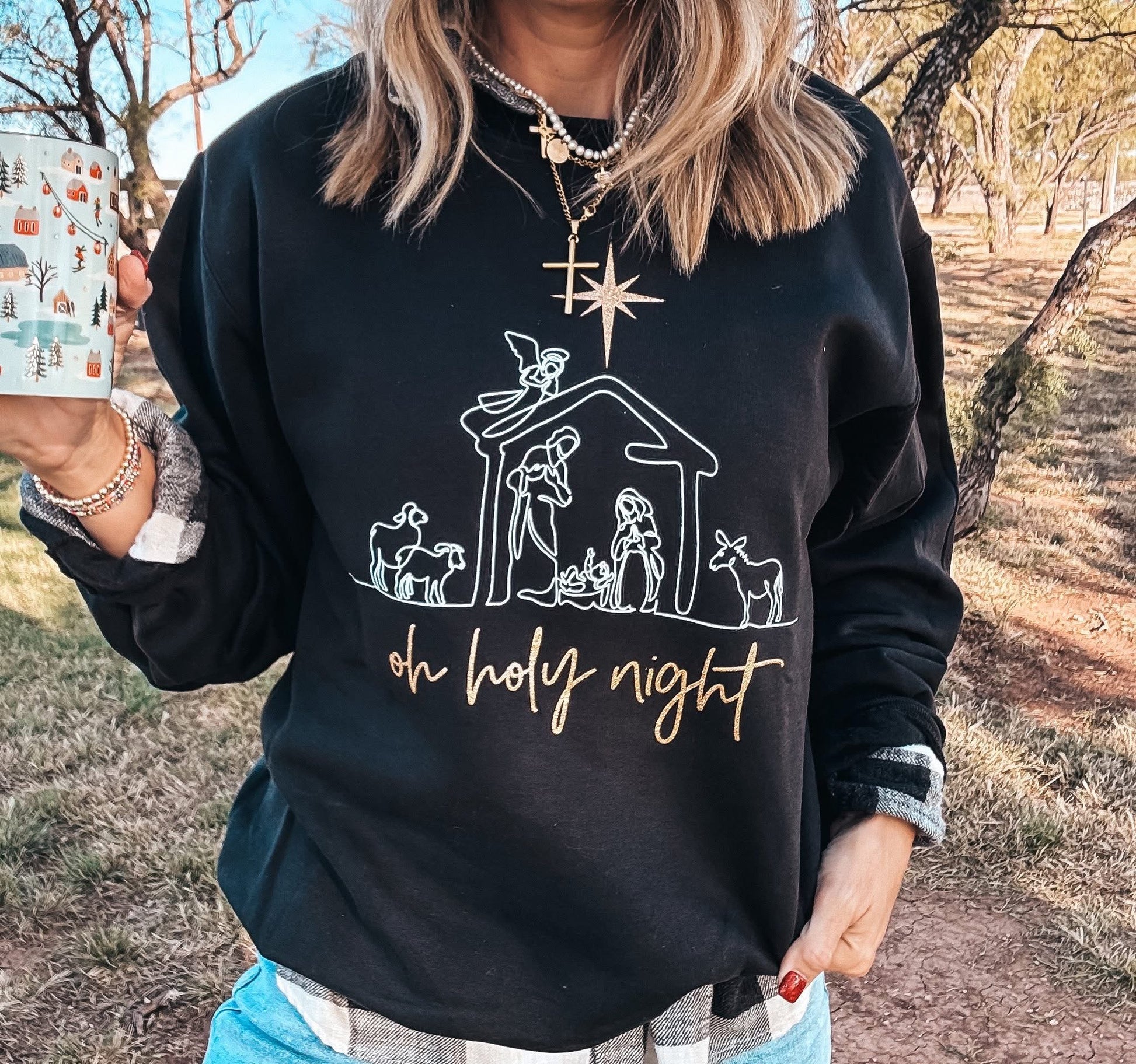 Person wearing a black sweatshirt with a nativity scene design and 'oh holy night' text, holding a mug outdoors.