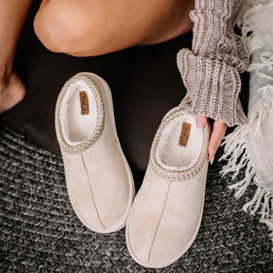 Beige slippers worn by a person sitting on a textured surface with a neutral sweater.