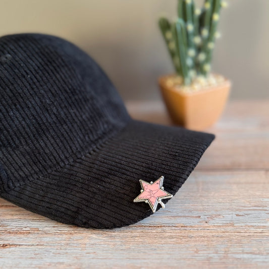 Black cap with a pink star pin on a wooden surface with a blurred cactus in the background