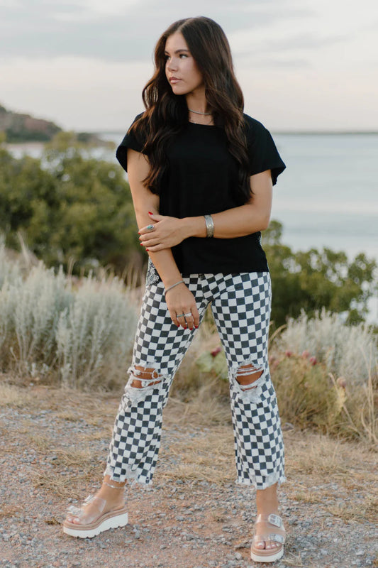Woman wearing a black top and checkered pants standing outdoors with a scenic background.
